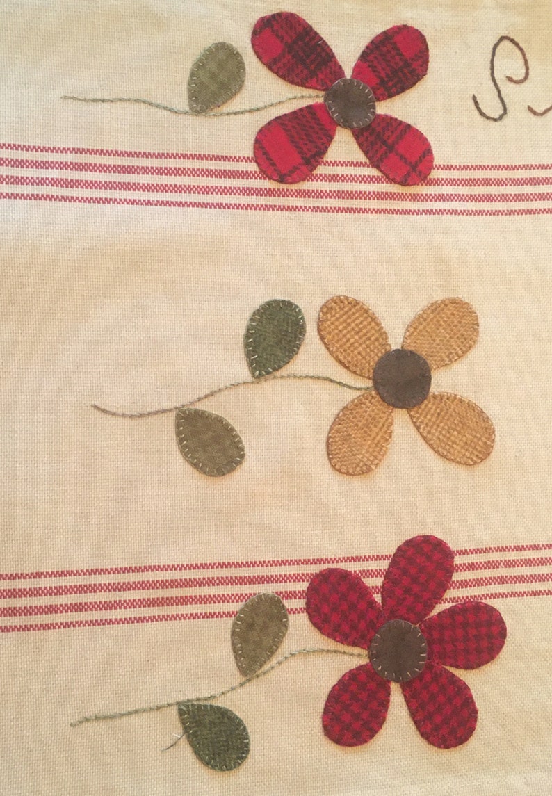 May include: A beige fabric kitchen towel with red and brown striped borders. Three appliqued flowers are stitched on the towel. The flowers are red, brown, and tan. The flowers have green leaves.