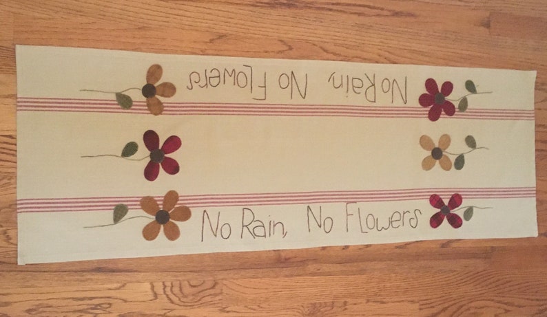 May include: A beige table runner with red and white stripes features the text "No Rain, No Flowers" repeated twice. The runner is decorated with embroidered flowers in shades of red, brown, and yellow.