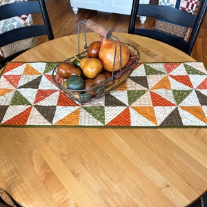 May include: A rectangular quilted table runner featuring a geometric pattern of triangles in shades of orange, green, brown, and white. A wire basket with a wooden handle sits on top, filled with various smooth, rounded decorative gourds.