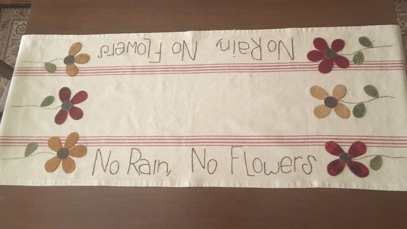May include: A white table runner with red stripes and the text "No Rain, No Flowers" embroidered in black. The runner is decorated with four felt flowers in red, yellow, and plaid.