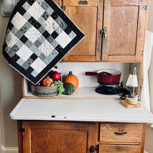 May include: A vintage wooden cabinet with a white countertop, displaying a black and white patchwork quilt, a bowl of fruit, and a small pumpkin. A red fondue pot sits on the counter, alongside a miniature lamp and a jar.