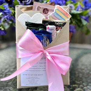 May include: A gift box wrapped in brown paper and tied with a pink satin ribbon. The box contains various items, including a sticker of books, heart-shaped items, and chocolate. The background features blue flowers.
