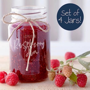May include: A glass jar filled with homemade raspberry jam, tied with twine and labeled "Homemade Raspberry Jam". The jar is sitting on a wooden surface with fresh raspberries and a sprig of raspberry leaves.