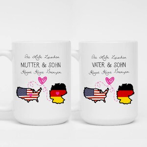 May include: Two white ceramic mugs with handles. One mug has the text "MUTTER & SOHN" and the other has "VATER & SOHN". Both mugs feature a graphic of the USA and German flags connected by a dotted line and a heart.
