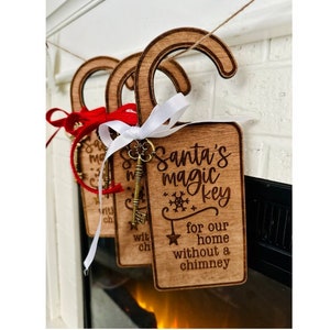 Puede incluir: Adornos navideños de madera con el texto "Santa's magic key" y una llave decorativa adjunta. Los adornos son rectangulares con una parte superior curvada y están atados con cintas rojas y blancas. También se incluye el texto "for our home without a chimney".