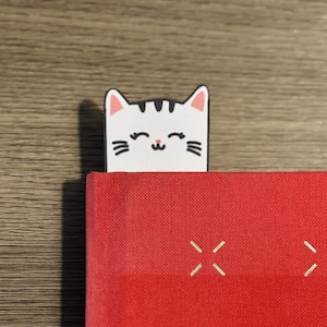 Puede incluir: Un marcapáginas blanco con forma de gato asoma por encima de un libro rojo. El marcapáginas tiene rayas y bigotes negros. El libro tiene texto blanco que dice "A Guide for Grown-".