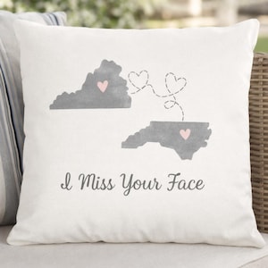 May include: White throw pillow with a gray map of two states, each with a pink heart. Dotted lines connect the states with three hearts. The text "I Miss Your Face" is printed below the maps.