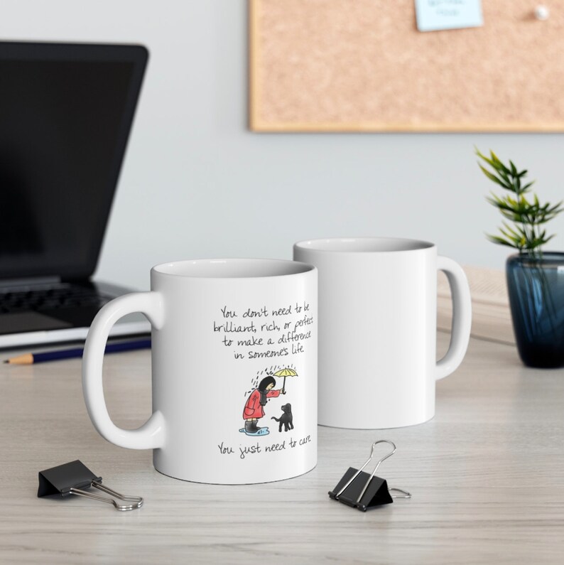 May include: White ceramic mug with a black and white illustration of a person holding an umbrella over a dog. The text on the mug reads "You don't need to be brilliant, rich, or perfect to make a difference in someone's life. You just need to care."