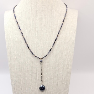 Könnte beinhalten: Eine zarte Halskette mit einer Kette aus kleinen, dunkelblauen Perlen mit goldfarbenen Akzenten. Ein vertikaler, silberfarbener Stab hält einen tropfenförmigen, dunkelblauen Anhänger. Die Halskette wird auf einer neutralfarbenen Schaufensterpuppe präsentiert.