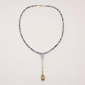 Könnte beinhalten: Eine zarte Halskette mit kleinen, runden, blau-grauen Perlen. Ein goldener Verschluss sichert die Kette. Ein silberner Anhänger hängt daran, verziert mit einem lila Edelstein und einem braun-gelben Edelstein.