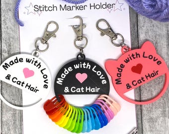 May include: Three stitch marker holders with the text "Made with Love & Cat Hair". One is white, one is black, and one is pink cat-shaped. A rainbow of stitch markers and a crochet hook are also visible.