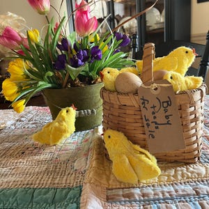 May include: A spring-themed still life featuring a woven basket filled with yellow chick figurines and eggs, with a tag that reads "Baby peeps for sale." A floral arrangement of pink and yellow tulips and purple irises sits nearby on a patchwork quilt.