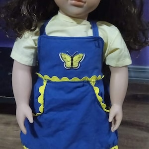 May include: A doll with brown hair and eyes, wearing a yellow shirt and a blue apron. The apron has a yellow butterfly design and yellow scalloped trim. The doll is standing.