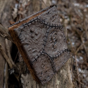 May include: A weathered, brown book with a textured cover and black stitching. The book's cover has a detailed, almost leathery appearance, with a central design and stitched edges. The book is resting on a tree branch, with a blurred natural background.