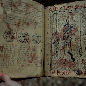 May include: Open book with aged, stained pages. The left page features text and circular symbols, while the right page displays an illustration of hanging figures with the text "Shatter Their Bones" and "They Deserve to Die".