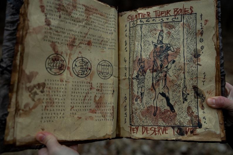 May include: Open book with aged, stained pages. The left page features text and circular symbols, while the right page displays an illustration of hanging figures with the text "Shatter Their Bones" and "They Deserve to Die".
