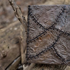May include: A handmade, textured book with a distressed, weathered appearance. The cover is a mottled gray with black stitching in a cross-stitch pattern. The book is resting on a weathered wooden surface.