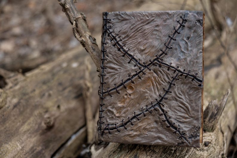 May include: A handmade, textured book with a distressed, weathered appearance. The cover is a mottled gray with black stitching in a cross-stitch pattern. The book is resting on a weathered wooden surface.