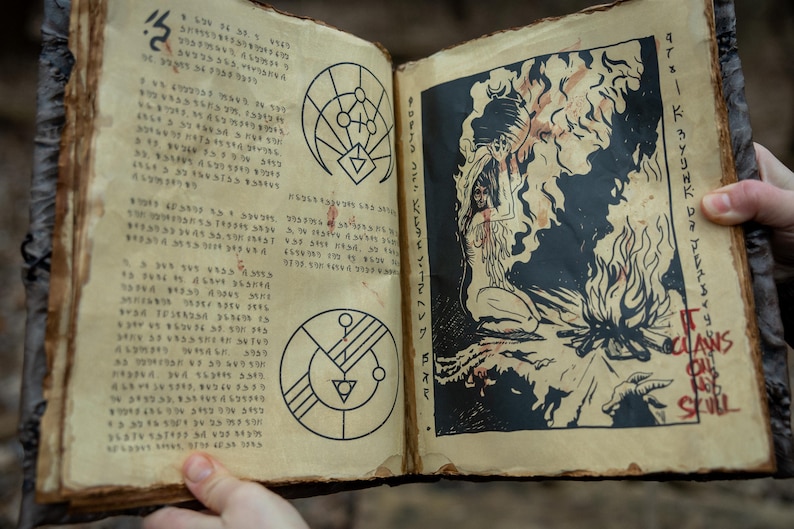 May include: Open book with aged, textured pages. The left page features handwritten text and geometric symbols. The right page displays a black and white illustration of a figure with flames and the words "IT CLAWS ON THE SKULL".