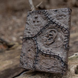 May include: A detailed, textured, brown book with a stitched design and an ear-shaped relief. The book's cover is intricately detailed with a dark stitch pattern. The book is resting on a weathered wooden surface.