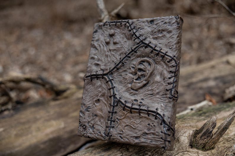 May include: A detailed, textured, brown book with a stitched design and an ear-shaped relief. The book's cover is intricately detailed with a dark stitch pattern. The book is resting on a weathered wooden surface.