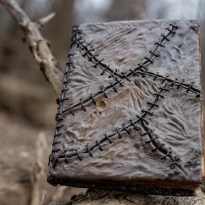 May include: A weathered, gray book with a textured cover and black stitching in a cross pattern. The book has a worn appearance, suggesting age or a vintage aesthetic. The spine and edges are visible, adding to the rustic charm.