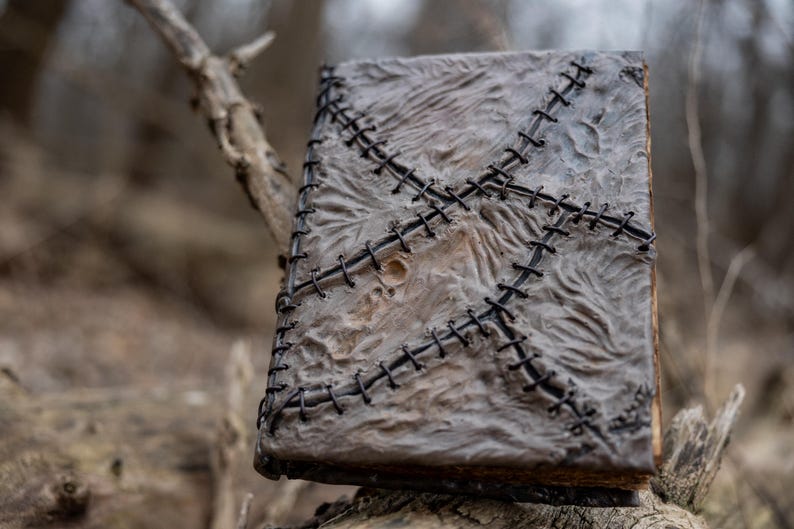 May include: A weathered, gray book with a textured cover and black stitching in a cross pattern. The book has a worn appearance, suggesting age or a vintage aesthetic. The spine and edges are visible, adding to the rustic charm.