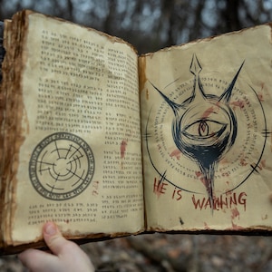 May include: An open, aged book with parchment-like pages. The left page features a circular design, while the right page displays an eye symbol with the text "HE IS WAITING" in red. The book has a dark, textured cover.