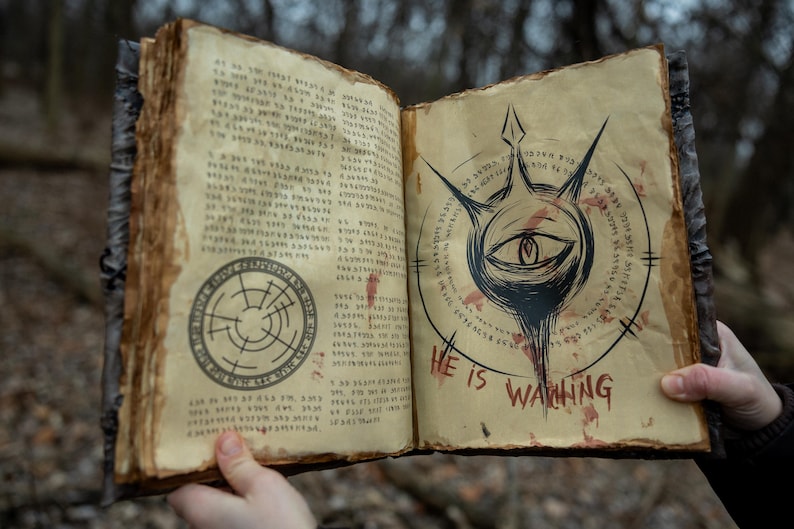 May include: An open, aged book with parchment-like pages. The left page features a circular design, while the right page displays an eye symbol with the text "HE IS WAITING" in red. The book has a dark, textured cover.