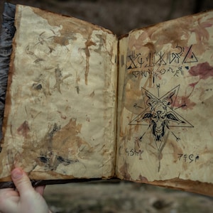May include: An open, aged book with stained pages. The right page features a pentagram with a goat's head and cryptic symbols. The left page has faded ink drawings. The book's cover is dark and textured, suggesting an antique or occult theme.
