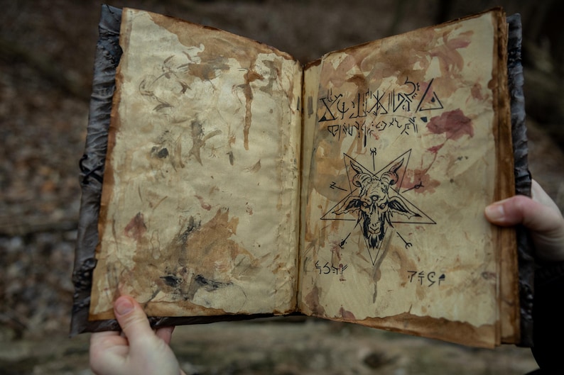 May include: An open, aged book with stained pages. The right page features a pentagram with a goat's head and cryptic symbols. The left page has faded ink drawings. The book's cover is dark and textured, suggesting an antique or occult theme.