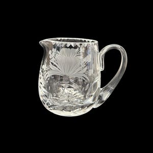 May include: A clear glass pitcher with a floral design etched into the side. The pitcher has a rounded body and a handle.