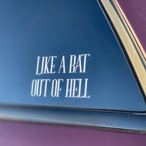 May include: A white vinyl decal applied to a dark car window, featuring the text "LIKE A BAT OUT OF HELL" in a tall, stylized, serif font. The lettering is arranged in two lines, with the words clearly visible against the dark glass surface.