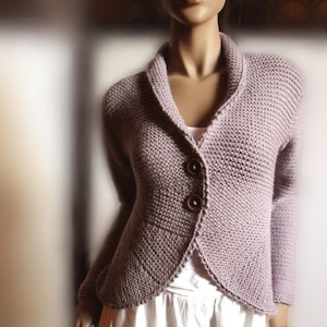 Könnte beinhalten: Eine lavendelfarbene Strickjacke mit Schalkragen, langen Ärmeln und zwei großen dunkelbraunen Knöpfen in der Taille. Das Kleidungsstück hat ein strukturiertes Strickmuster und einen geschwungenen Saum und wird über einem weißen Rock getragen.