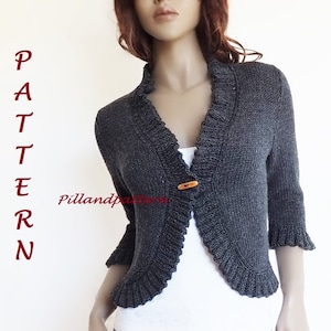 Könnte beinhalten: Ein grauer Strickcardigan mit Rüschen an Ausschnitt und Ärmeln. Der Cardigan hat einen einzigen Knopfverschluss und ist taillenkurz.