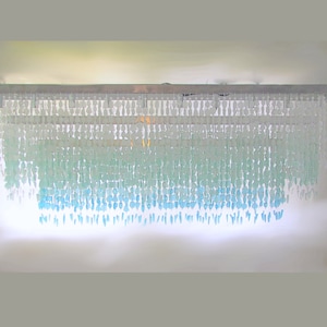 Puede incluir: Una lámpara de araña moderna de varios niveles con un diseño en cascada. La lámpara de araña está hecha de cuentas de vidrio transparentes que están graduadas en tamaño, creando un efecto de capas. Las cuentas están dispuestas en un degradado de colores, comenzando con blanco en la parte superior y pasando a azul claro en la parte inferior.