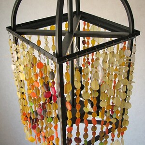 Puede incluir: Una l&aacute;mpara de ara&ntilde;a de metal negro con un marco cuadrado y m&uacute;ltiples hebras de cuentas de vidrio de colores que cuelgan de ella. Las cuentas son en tonos de amarillo, naranja, rojo y verde.