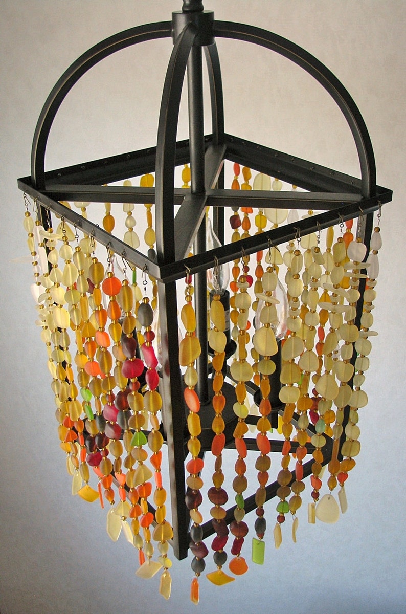 Puede incluir: Una l&aacute;mpara de ara&ntilde;a de metal negro con un marco cuadrado y m&uacute;ltiples hebras de cuentas de vidrio de colores que cuelgan de ella. Las cuentas son en tonos de amarillo, naranja, rojo y verde.