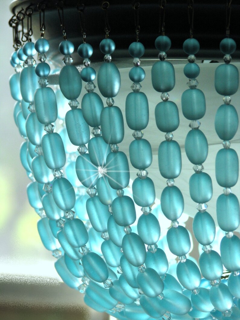 May include: A chandelier made of light blue glass beads. The beads are oval shaped and hang in rows. The chandelier is lit from behind, creating a sparkling effect.