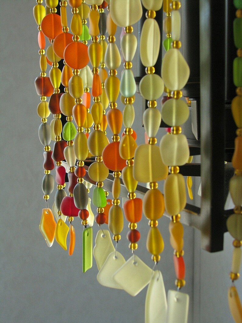 Puede incluir: Una cortina de cuentas hecha de cuentas de vidrio en varios colores, incluyendo naranja, amarillo, verde y blanco. Las cuentas est&aacute;n enhebradas en alambre y cuelgan de un marco de metal negro.