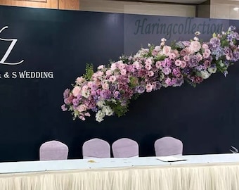 Pode incluir: Um cenário de cerimônia de casamento com fundo preto e uma guirlanda floral branca com flores roxas e rosas. O texto "A & S Wedding" está no cenário. Há uma mesa branca com uma toalha de renda branca e quatro cadeiras com almofadas roxas.