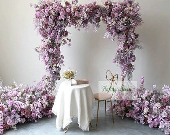 Pode incluir: Uma mesa branca com uma toalha branca está posta com um livro e um pequeno buquê de flores. Uma cadeira dourada com encosto de laço está posicionada à direita da mesa. O fundo apresenta um grande arco floral feito de flores artificiais roxas e brancas.