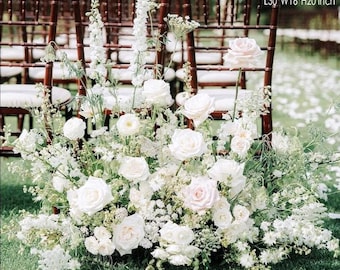 Op de afbeelding: Een weelderig bloemstuk met witte rozen en andere witte bloemen, geaccentueerd met groen. De opstelling is ongeveer 76 cm lang, 41 cm breed en 51 cm hoog. De bloemen staan tegen een achtergrond van houten stoelen.
