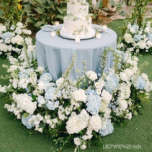 May include: A three-tiered white wedding cake with floral decorations sits on a light blue table. The table is surrounded by white and light blue flowers. The table is 80 inches long, 16 inches wide, and 20 inches high.