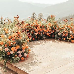 Puede incluir: Arreglo floral con flores de color naranja y melocotón y hojas verdes, dispuesto sobre una plataforma de madera. El arreglo mide aproximadamente 381 cm de largo, 41 cm de ancho y 51 cm de alto. Se ven montañas al fondo.