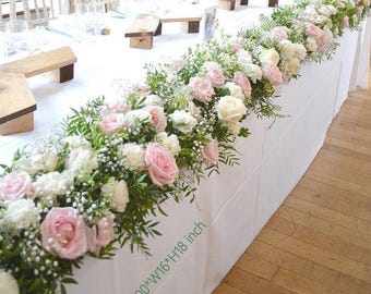 Puede incluir: Un mantel blanco adornado con un arreglo floral de rosas rosas y blancas, paniculata y vegetación. El arreglo mide aproximadamente 254 cm de largo, 41 cm de ancho y 46 cm de alto. La mesa está preparada para un evento.