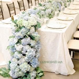 Könnte beinhalten: Ein langer weißer Tisch ist für eine formelle Veranstaltung gedeckt, geschmückt mit einem Läufer aus blauen und weißen Hortensien. Der Tisch ist mit einer weißen, gemusterten Tischdecke bedeckt. Abmessungen: L305*B41*H51 cm.