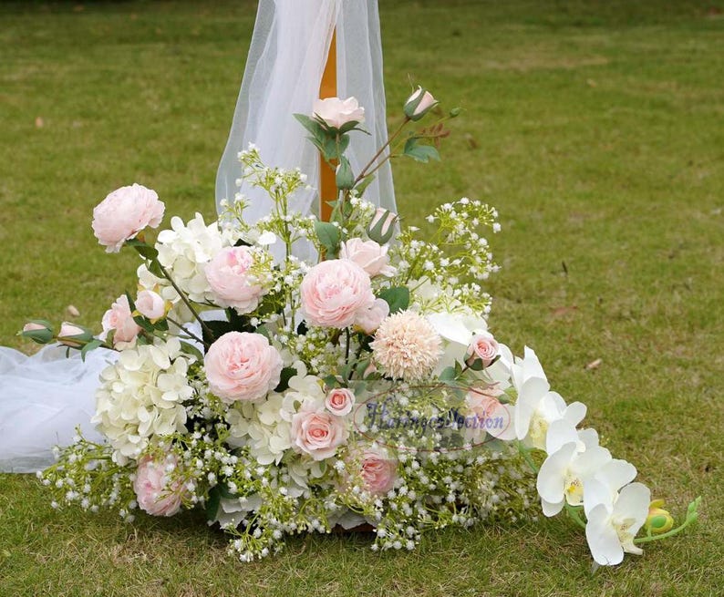 May include: A wedding arch decorated with pink and white flowers, including roses, hydrangeas, and baby's breath. White sheer fabric drapes over the arch.
