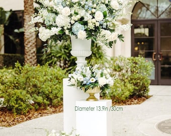 May include: Three floral arrangements featuring white and blue flowers. The largest arrangement is in a white urn, with a diameter of 60cm. The other two arrangements are in gold-coloured bowls, with diameters of 30cm and 36cm. The arrangements are on white pedestals.