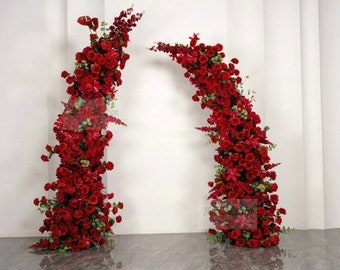 May include: Two tall floral arrangements made of red roses and greenery. The arrangements are in the shape of a column and an arch. The arrangements are against a white wall.